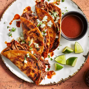 Three birria tacos onw a white round plate with a gravy bowl and lime slices.