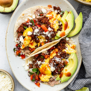 Two breakfast tacos in a round white plate with sliced avocados.